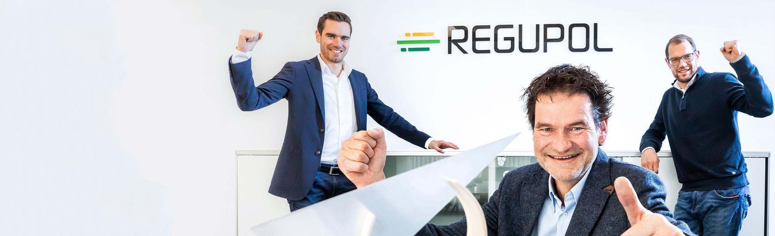 REGUPOL-CEOs mit Umweltwirtschaftspreis – Nachhaltigkeit, Auszeichnung, Erfolg Drei Männer, die CEOs von REGUPOL, stehen fröhlich und mit erhobenen Armen in einem hellen Raum vor dem Firmenlogo. Im Vordergrund steht ein Umweltwirtschaftspreis mit moderner, skulpturaler Form – Symbol für unternehmerische Nachhaltigkeit und Erfolg.