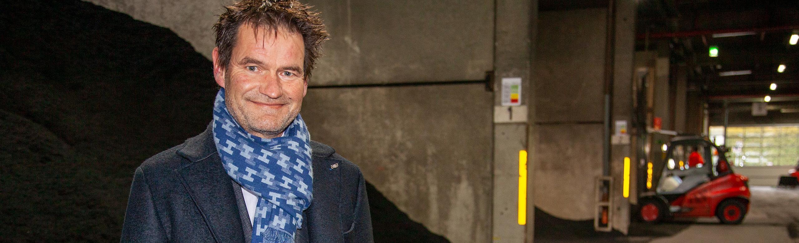 REGUPOL-CEO in Produktion – Recycling, Industrie, Führung, Umweltbewusstsein Ein CEO von REGUPOL steht in einer Industriehalle mit dunkler Kleidung und blau gemusterem Schal. Im Hintergrund sind große Materiallager und ein roter Gabelstapler im Einsatz zu sehen.