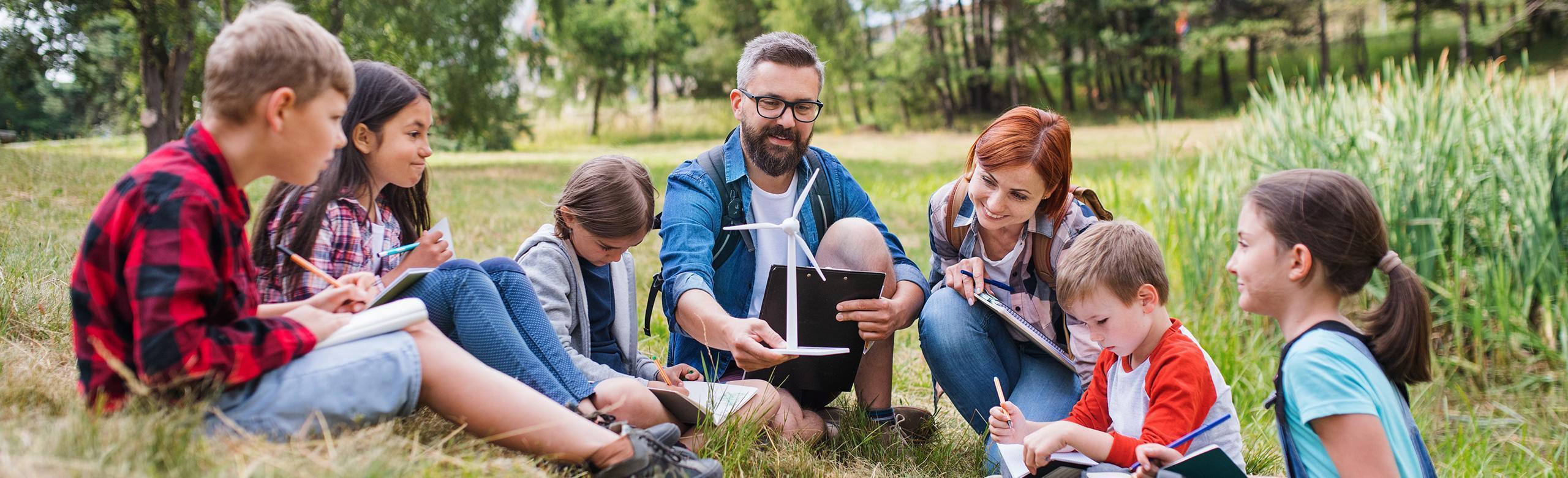 Eine Gruppe von Kindern sitzt mit zwei Erwachsenen im Kreis auf einer Wiese. Sie lernen gemeinsam im Freien und beschäftigen sich mit Notizbüchern und einem Modell eines Windrads. Im Hintergrund sieht man Bäume und Schilf.