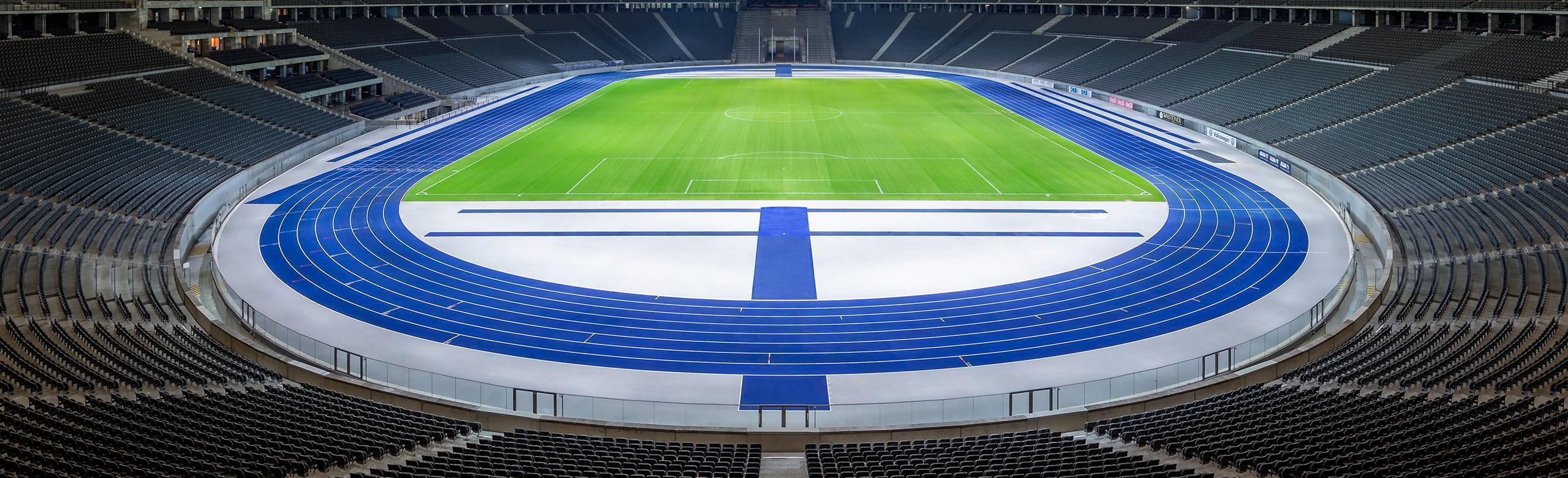 Blick ins leere Olympiastadion Berlin mit der markanten blauen REGUPOL-Laufbahn rund um das grüne Spielfeld. Die Tribünen sind leer,